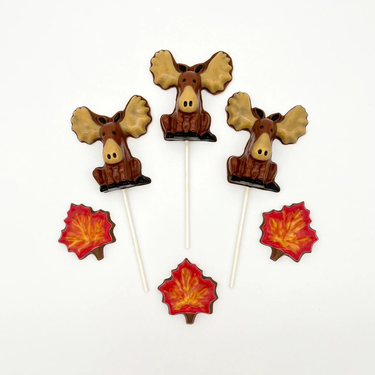 Front view of 3 chocolate brown moose suckers with big beige antlers and snouts, alongside colourful red chocolate maple leaves.