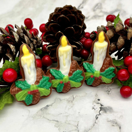 Chocolates in the shape of a candle, with a green ribbon, holly leaves, red berries and pinecones. Surrounded by festive decorations.