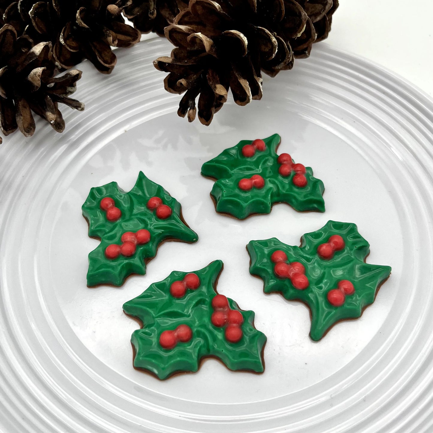 Green holly leaf chocolates with red berries on a white plate with pine cones.