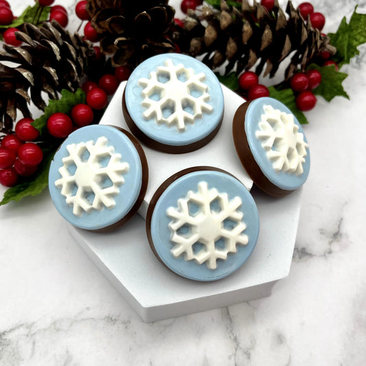 Four chocolate medallions with white and icy blue coloured snowflake designs. Displayed on a white stand, surrounded by pine cones and berries.
