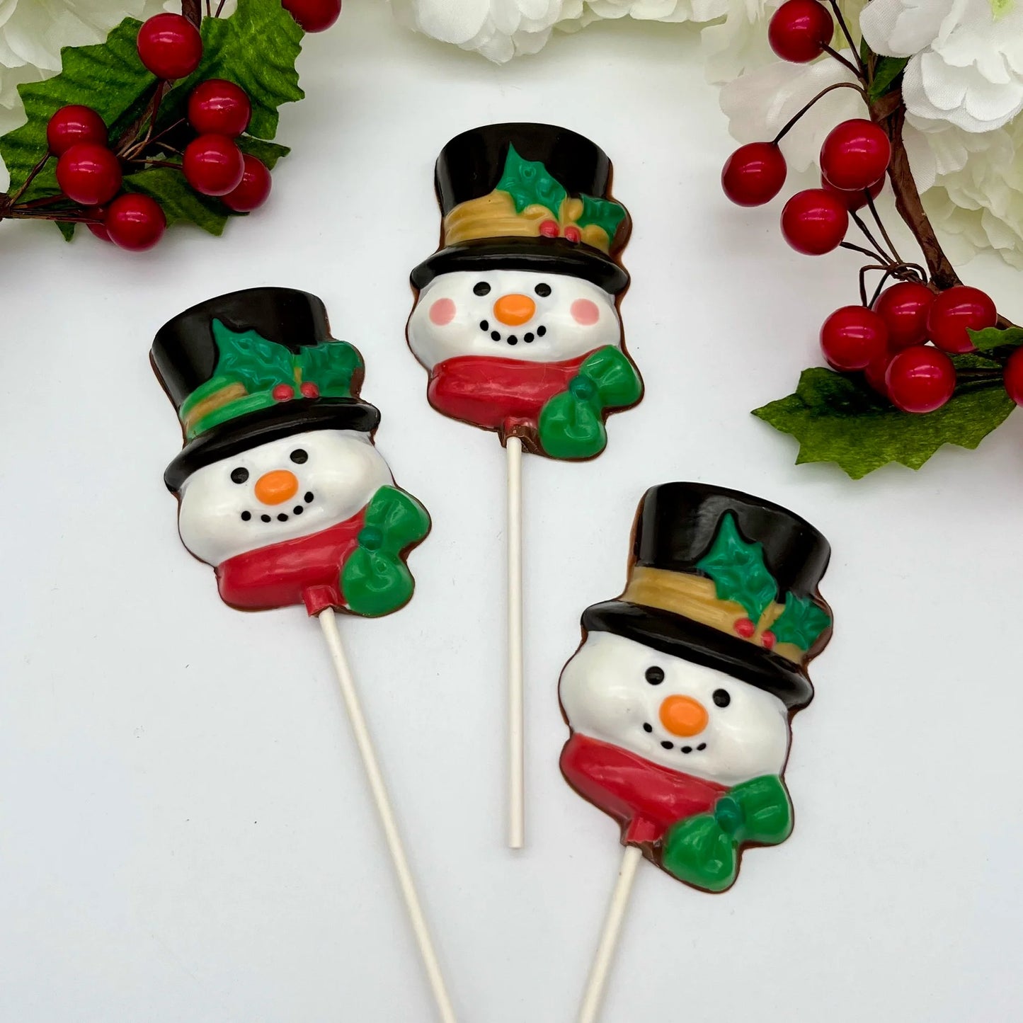 Chocolate snowman suckers wearing black top hats with holly and red scarves, surrounded by festive decorations.