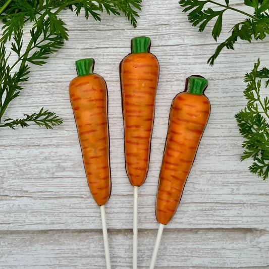 Realistic-looking carrot chocolate suckers with orange and green detailing, surrounded by leafy carrot greens on a wood background.