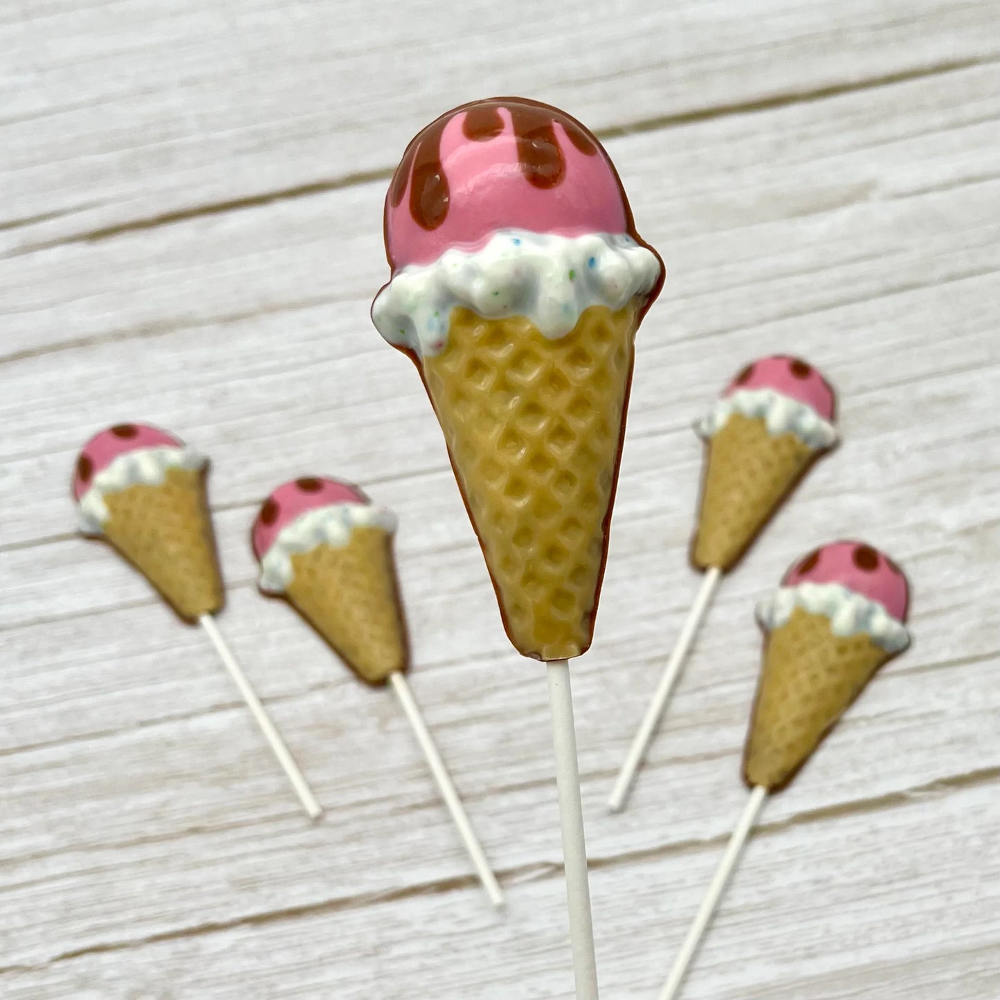 Ice cream cone chocolate suckers decorated in neopolitan colours; light brown cone, candy-sprinkled white, bright pink, topped with brown dripping like melting chocolate.