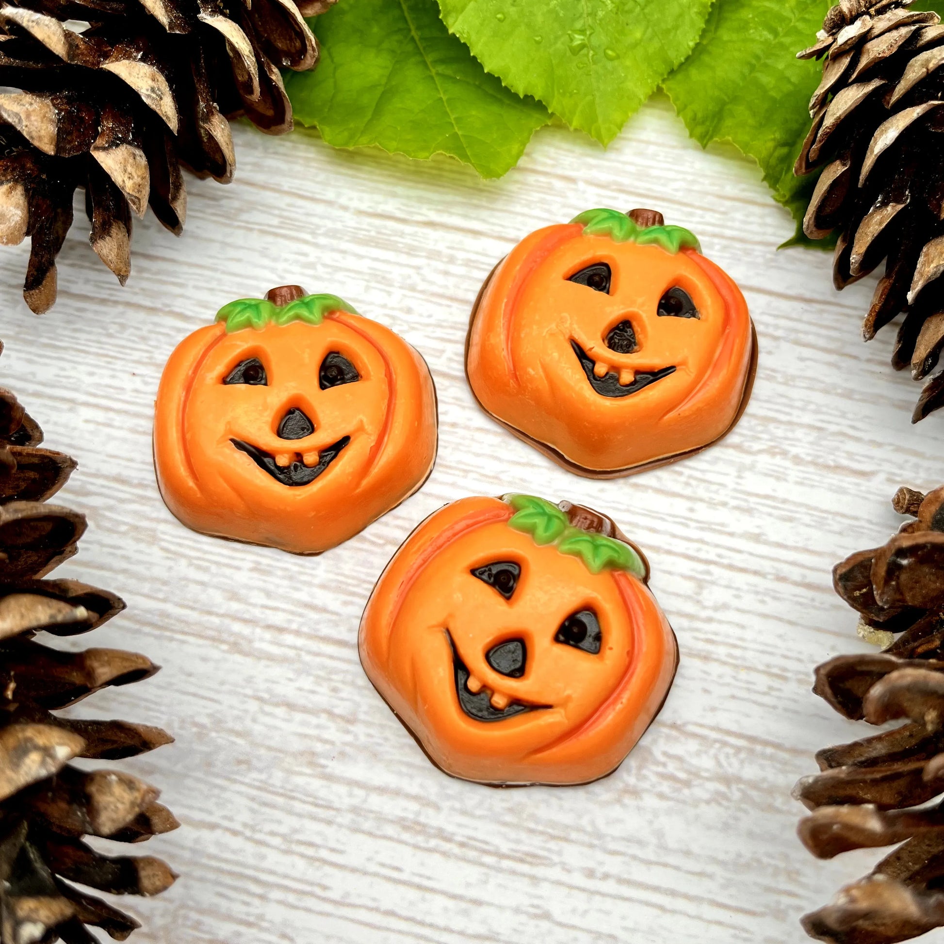Three orange chocolate pumpkins, "carved" with toothy grins, surrounded by pinecones and leaves on a light wooden surface.