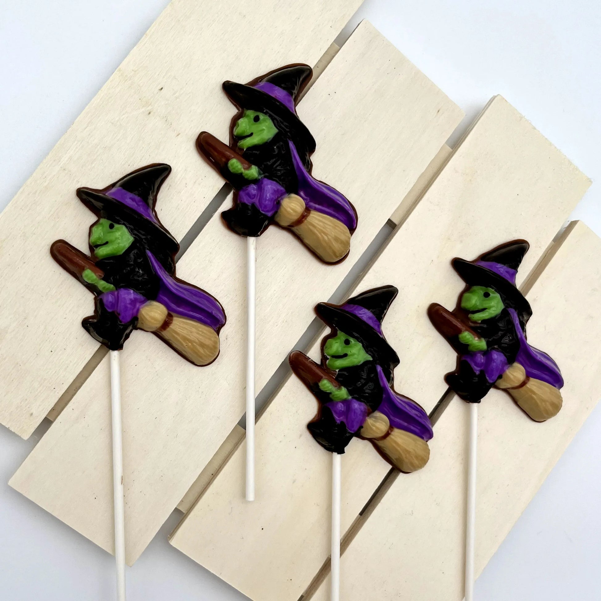 Chocolate suckers of witches, with green faces, purple capes, black hat and clothing, all flying on brooms; pictured on a wooden surface.