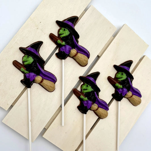 Chocolate suckers of witches, with green faces, purple capes, black hat and clothing, all flying on brooms; pictured on a wooden surface.