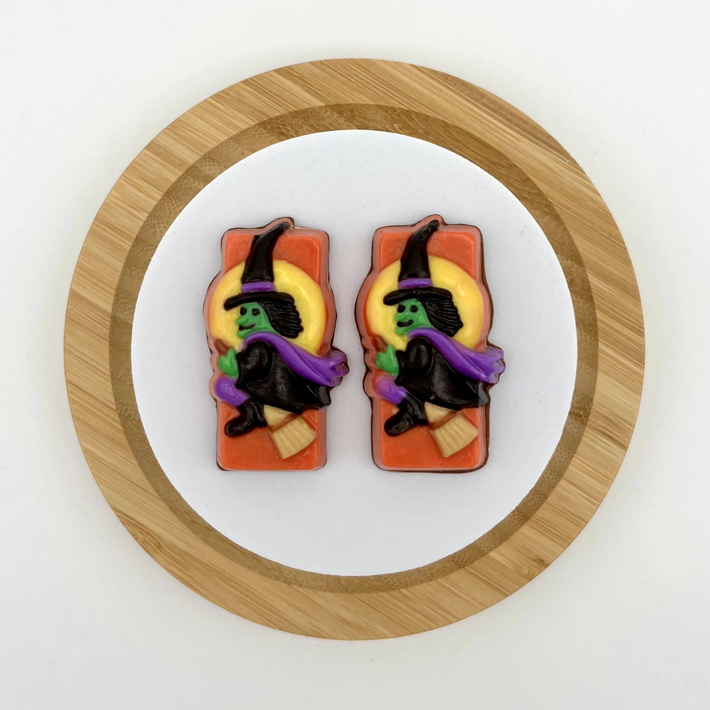 Classic Halloween witch in a flowing purple cape and green face aglow, soaring across a glowing full moon on an orange background, all made of chocolate.