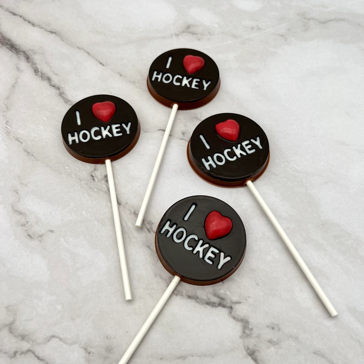 Four 'I ❤️ Hockey' chocolate suckers on a marble surface. White lettering and a red heart on a round black "puck" background.