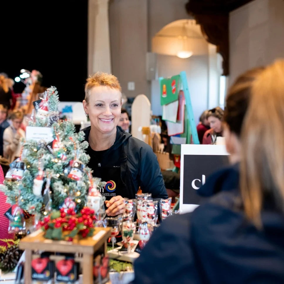 Owner at a craft fair with a small decorated Christmas tree and booth setup.