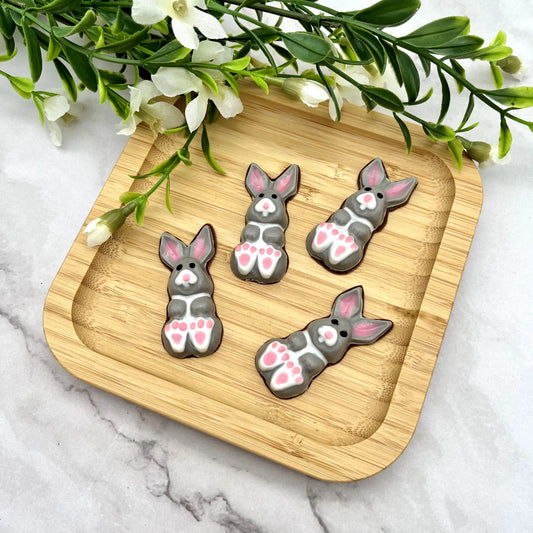 Grey and white chocolate bunnies with little pink toes and ears, displayed on a wooden tray with greenery in the background.