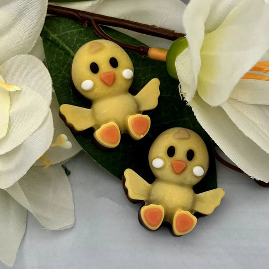 Two yellow baby chicks with orange feet and beaks made of chocolate. Pictured with green leaves and white flowers.