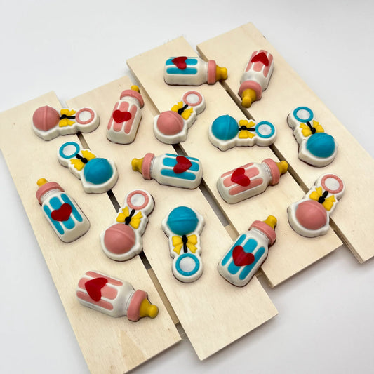 Multiple hand-painted chocolates of pink and blue-themed baby rattles with yellow ribbons, and milk bottles with red hearts.