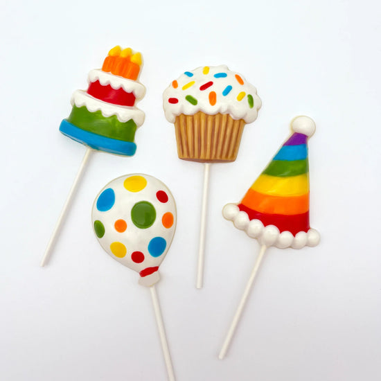 Four hand-painted chocolate suckers, shaped like a cake, cupcake, balloon, and party hat, coloured in a festive rainbow palette.