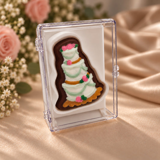 Chocolate tiered wedding cake decorated with pink roses and green garlands in a clear acrylic gift box.