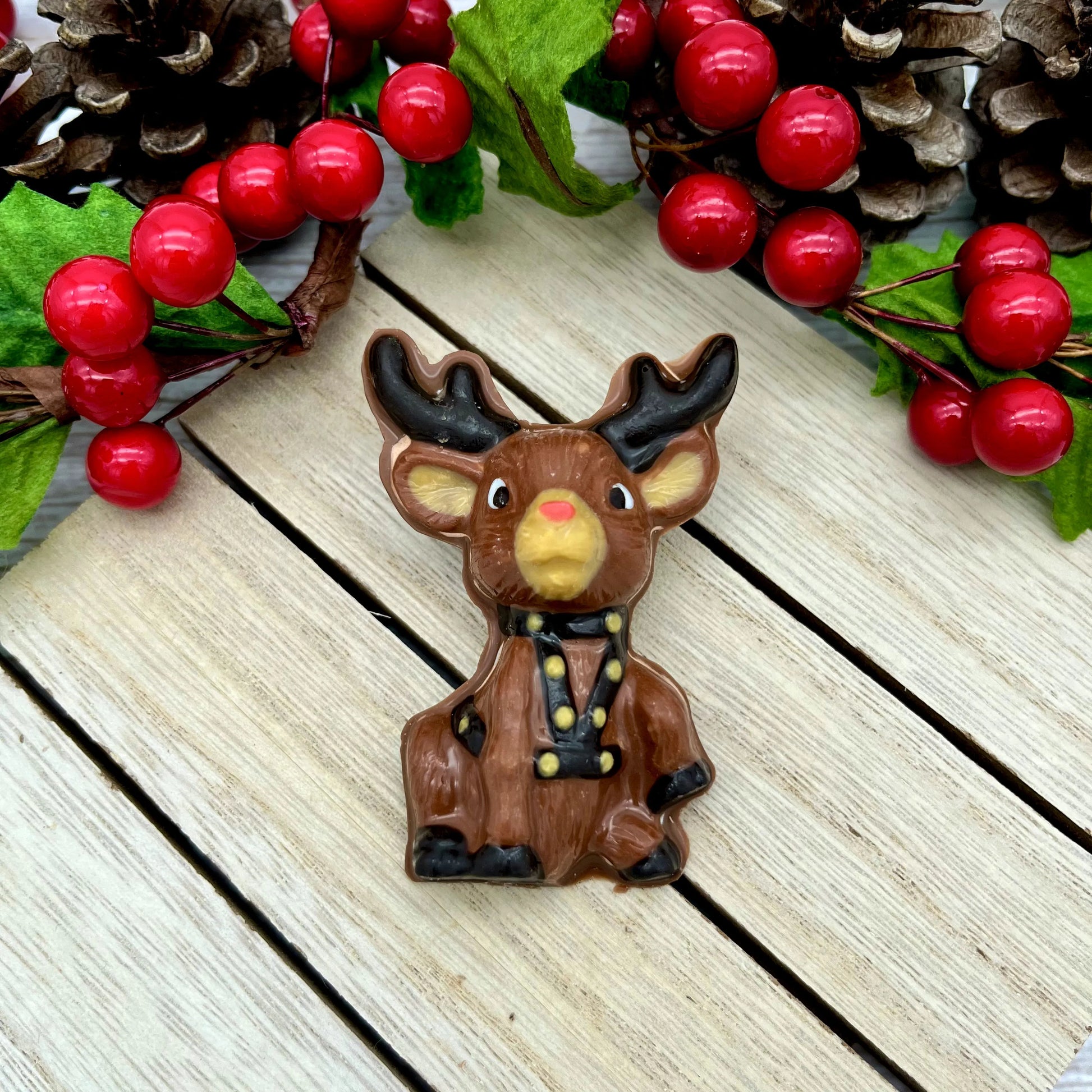 Brown chocolate reindeer, with red nose, dark antlers, harness with golden bells, surrounded by pinecones and berries on a wood surface.