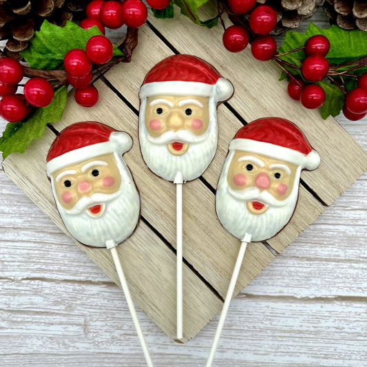 Three chocolate Santa suckers with red hats and rosy cheeks, arranged on a wooden board with festive berries and pinecones.