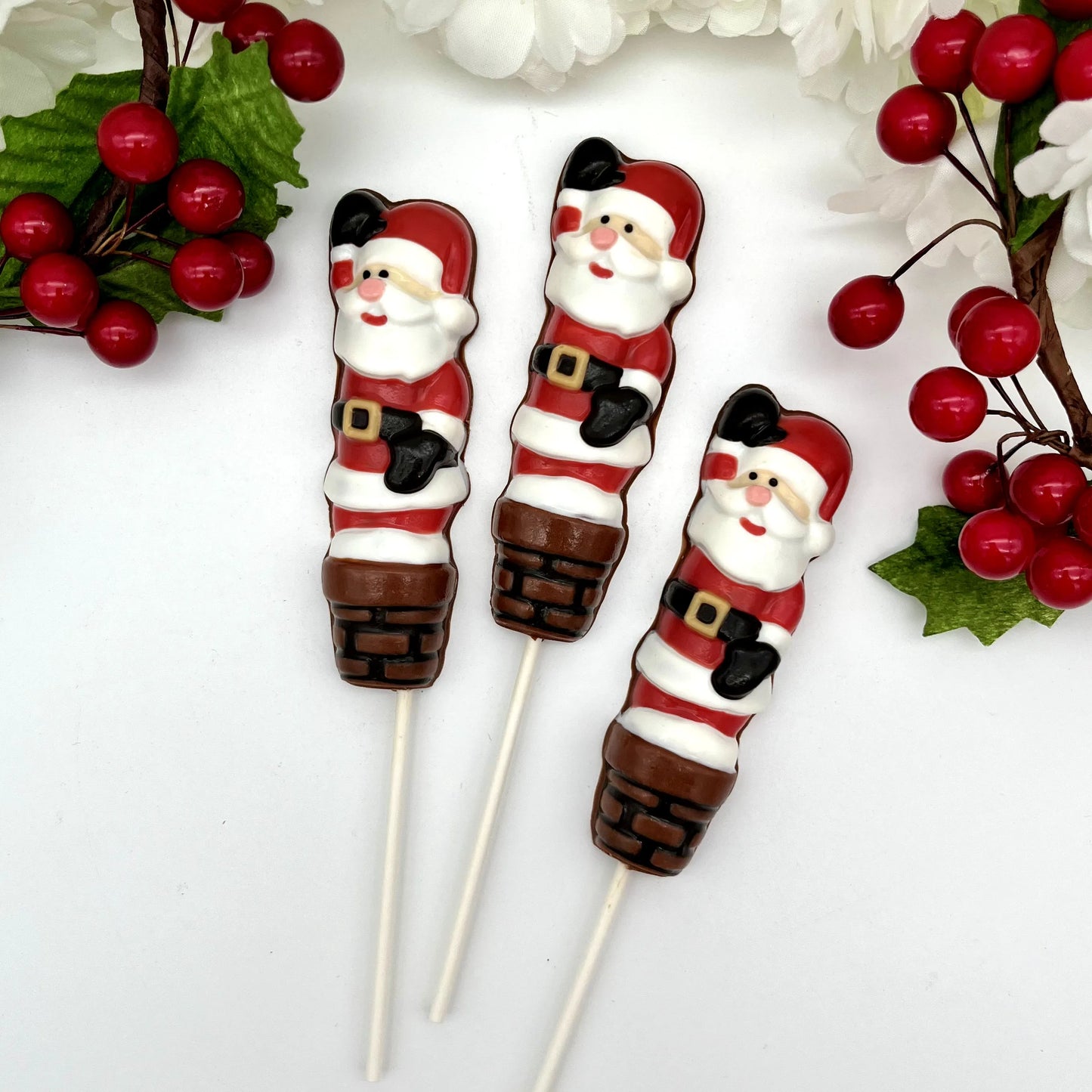Three chocolate suckers of Santa Claus going down a chimney, with Christmas decorations in the background.

Three chocolate suckers with Santa Claus design on a stick, against a white background.