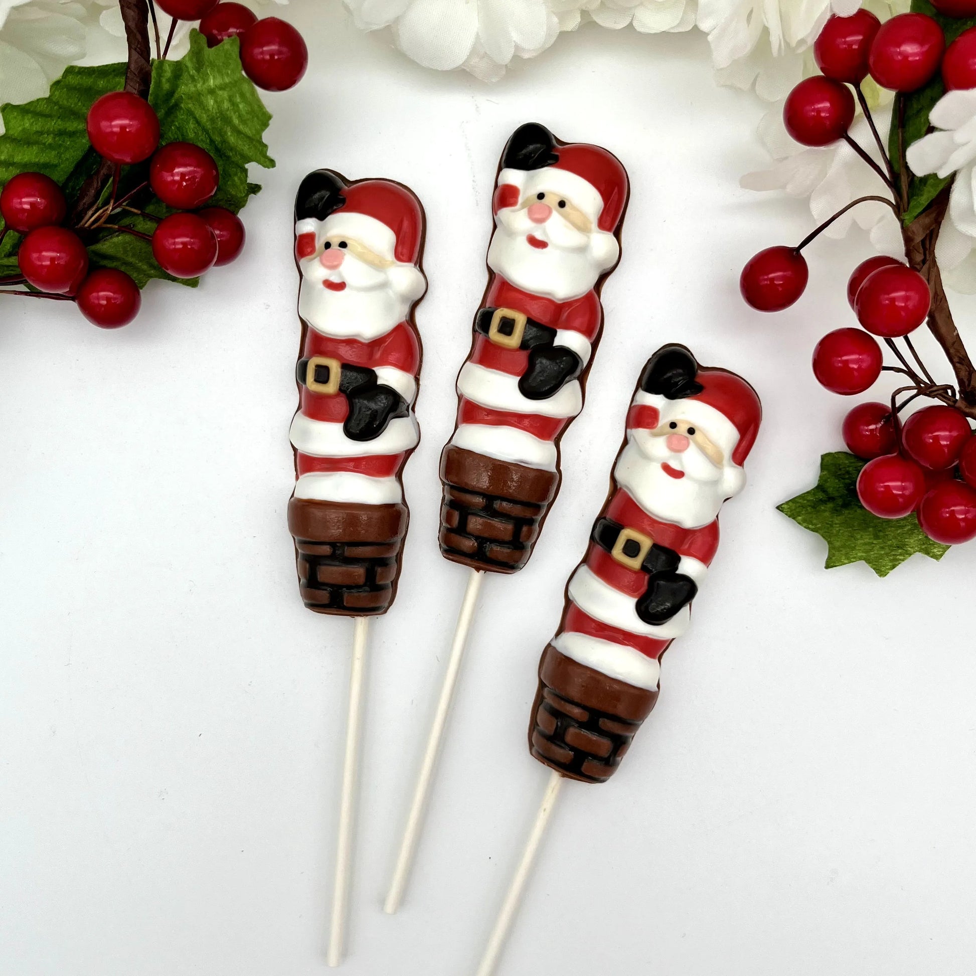 Three chocolate suckers of Santa Claus going down a chimney, with Christmas decorations in the background.

Three chocolate suckers with Santa Claus design on a stick, against a white background.