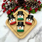 Four marshmallow-shaped snowman chocolates on a wooden board with Christmas decorations and marble table in the background.