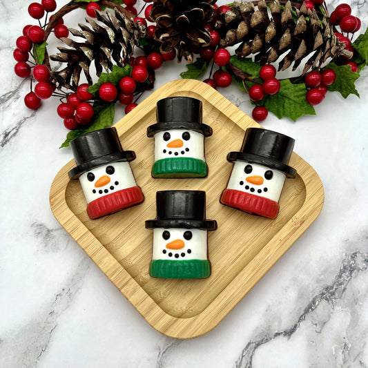 Four marshmallow-shaped snowman chocolates on a wooden board with Christmas decorations and marble table in the background.