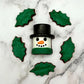 Marshmallow-shaped snowman chocolate with a black top hat and green scarf, surrounded by holly leaves on a marble surface.