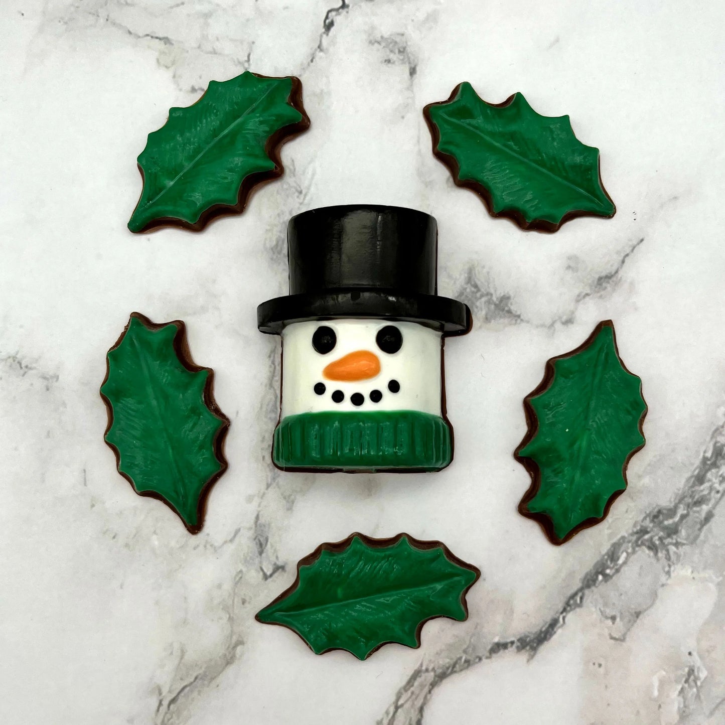 Marshmallow-shaped snowman chocolate with a black top hat and green scarf, surrounded by holly leaves on a marble surface.