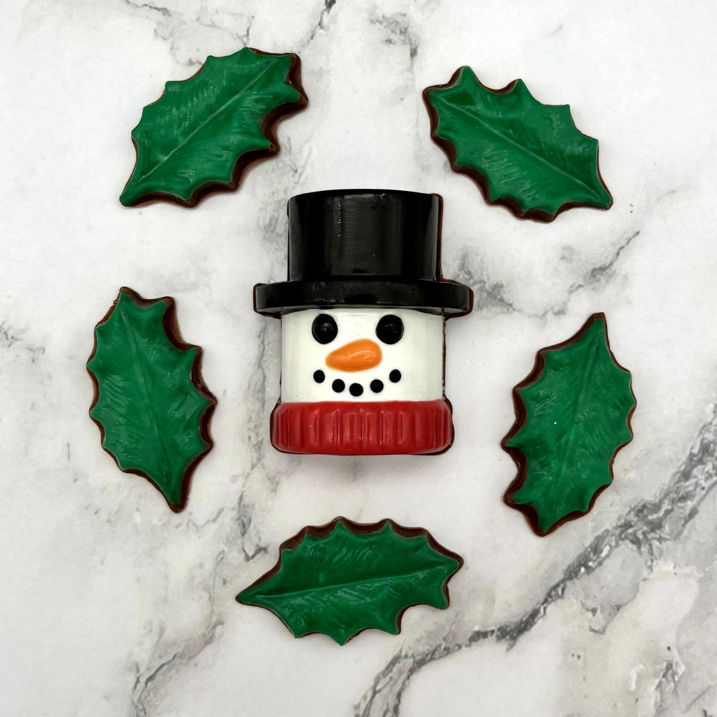 Marshmallow-shaped snowman chocolate with a black top hat and red scarf, surrounded by holly leaves on a marble surface.