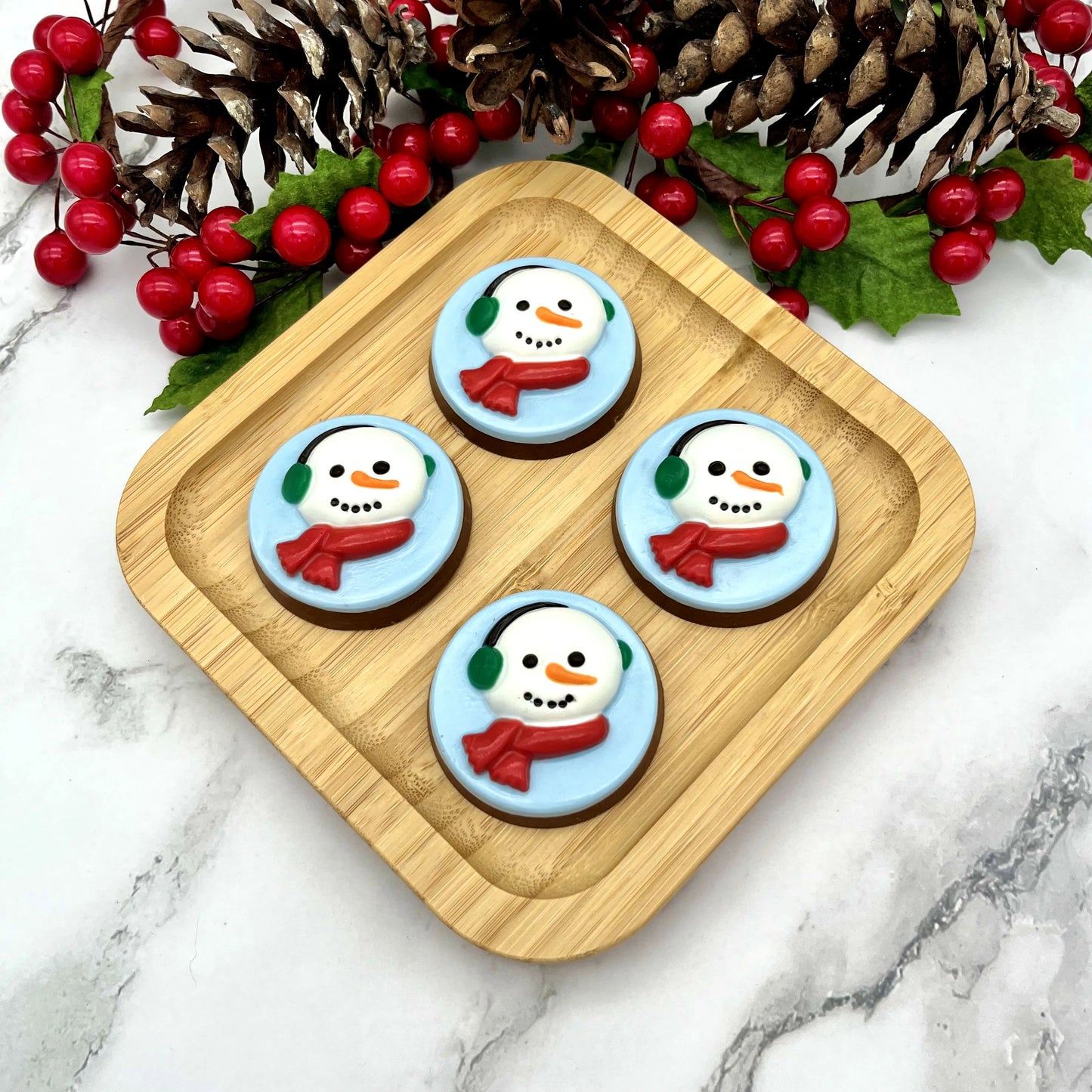 Four round chocolate medallions adorned with snowman faces on a blue background, with green ear muffs and red scarves. 