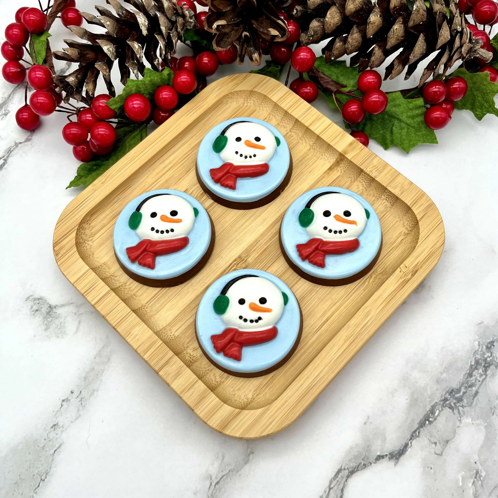 Four round chocolate medallions adorned with snowman faces on a blue background, with green ear muffs and red scarves. 