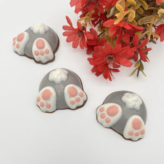 Three fun grey-coloured chocolate bunny butts with little pink toes and white tails. Red flowers create a festive springtime scene.