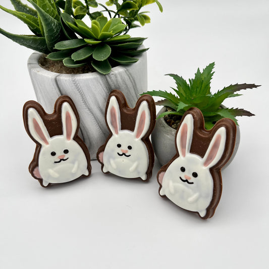 Three white and pink emoji-style bunny chocolates, propped up on little green planters.