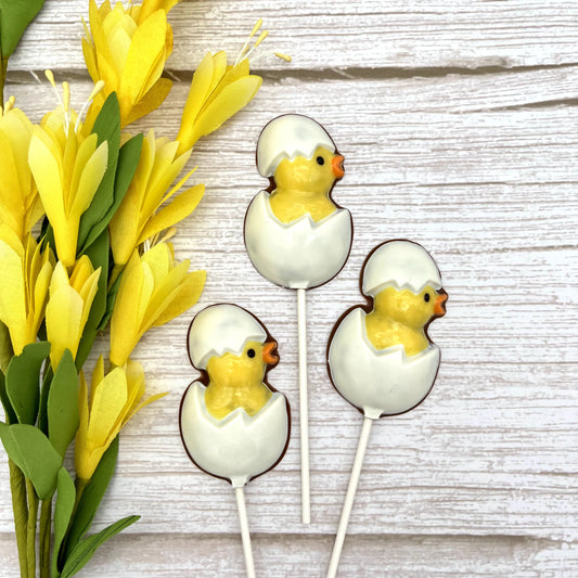 A chocolate sucker designed to look like a chick inside an egg cracked open, displayed on a wood surface with yellow flowers.