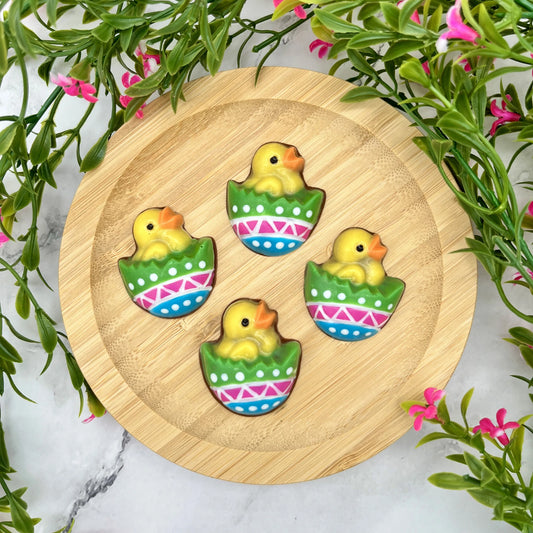 Hand-painted chocolates of yellow chicks in colourful eggs, decorated with dots and zigzags, arranged on a plate with spring flowers.
