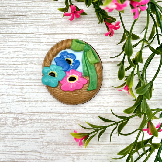A chocolate basket with a green ribbon, with three Forget-me-not flowers in shades of blue, pink and turquoise.