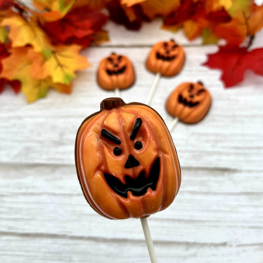 Pumpkin-shaped chocolate on a stick, with wicked jack-o'-lantern face. Surrounded by small pumpkin decorations and autumn leaves.