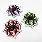 Three jumbo black widow spider chocolates, painted with red eyes and hourglass marking, white web on red, purple and green backgrounds.