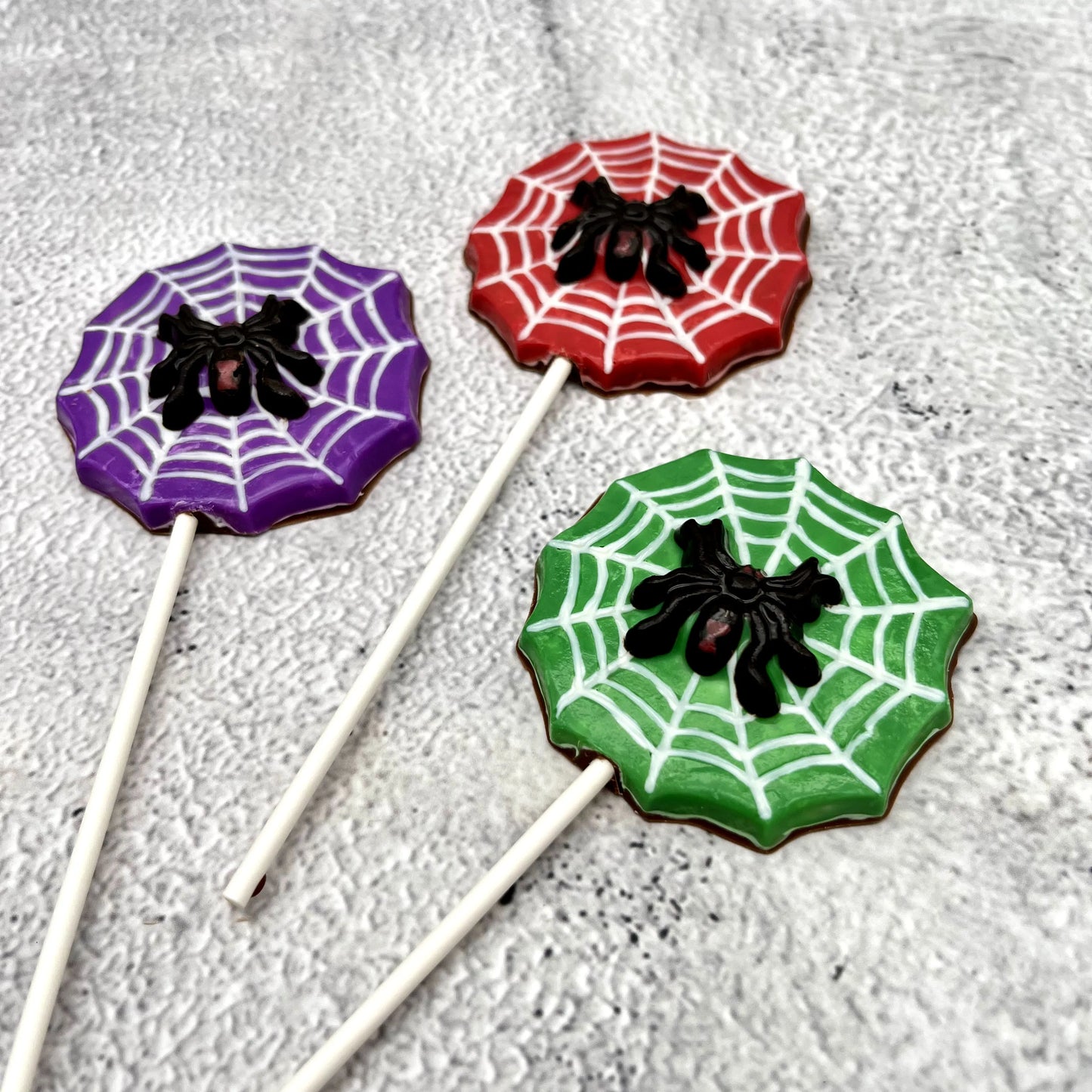 Black widow spider atop a white web, on a red, green or purple background shaped like a web. All made of chocolate as suckers.