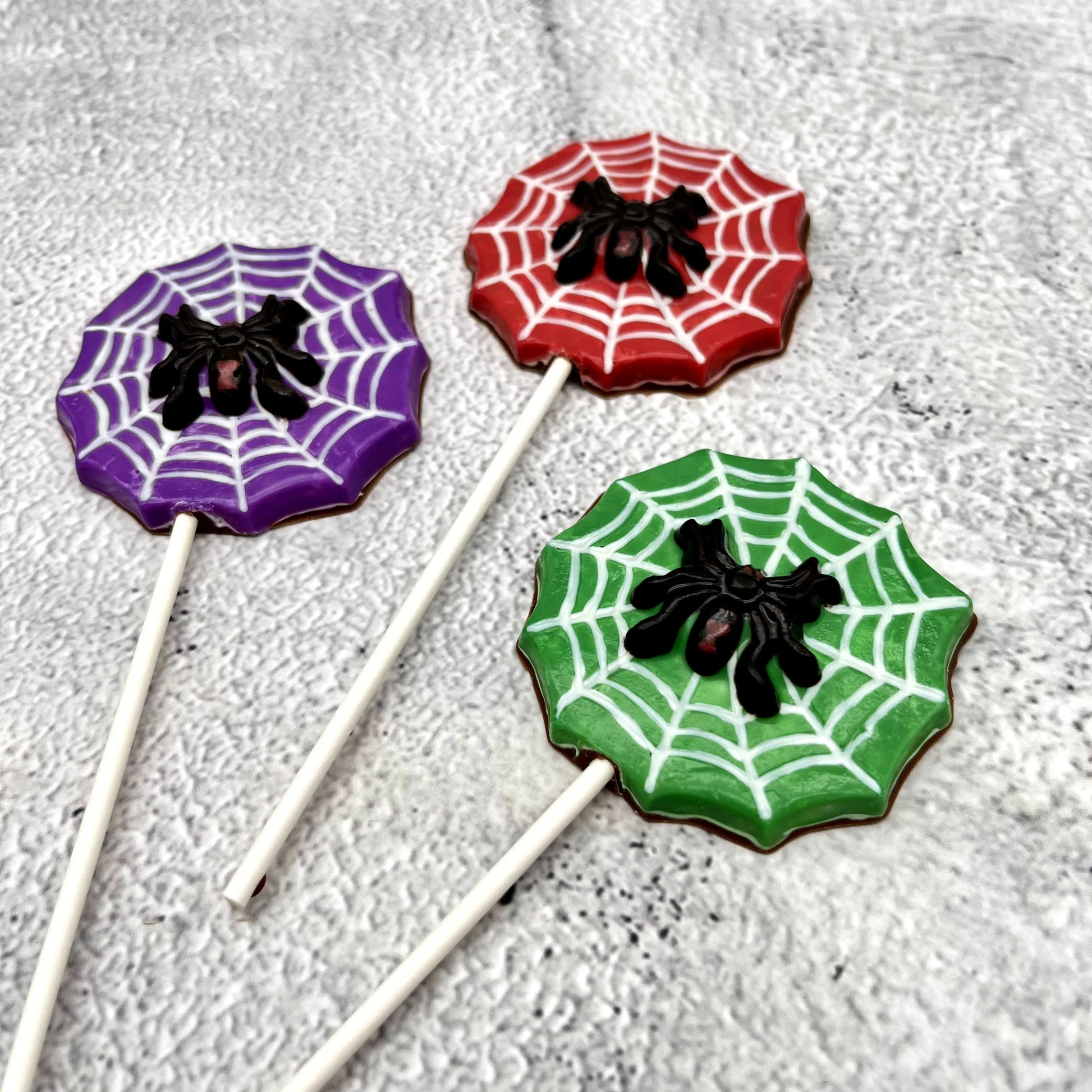 Black widow spider atop a white web, on a red, green or purple background shaped like a web. All made of chocolate as suckers.