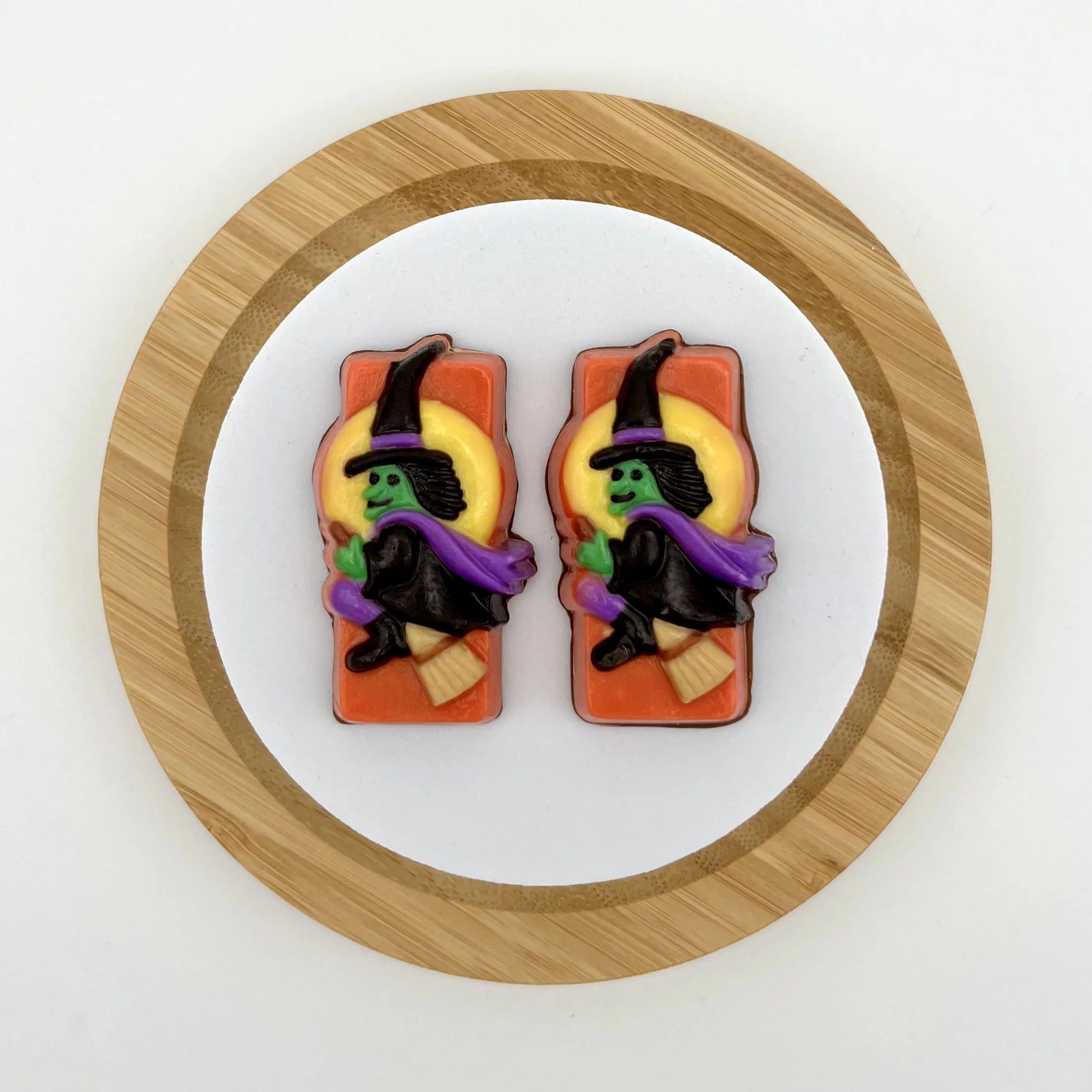 Classic Halloween witch in a flowing purple cape and green face aglow, soaring across a glowing full moon on an orange background, all made of chocolate.