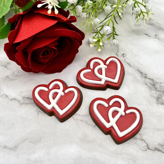 Three red heart-shaped chocolates with white detail, placed on a marble surface with red rose decor.