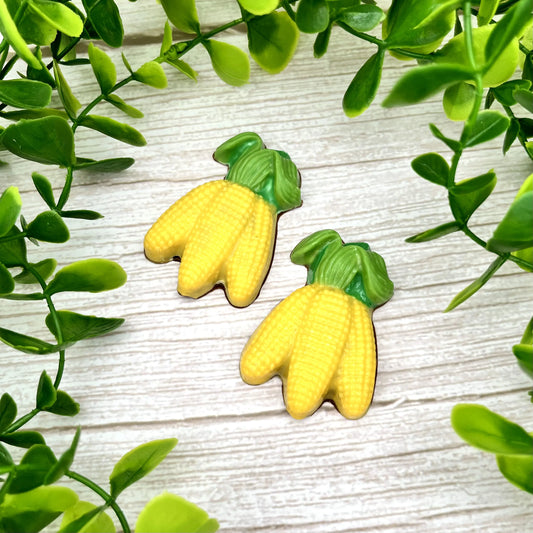 Two yellow corn-shaped chocolates with green leaves, pictured on a light wooden surface surrounded by green leaves.