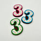 Three colourful number '3' chocolates, hand-painted with multi-colour dots and sprinkles. Displayed on a white background.