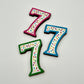 Three colourful number '7' chocolates, hand-painted with multi-colour dots and sprinkles. Displayed on a white background.