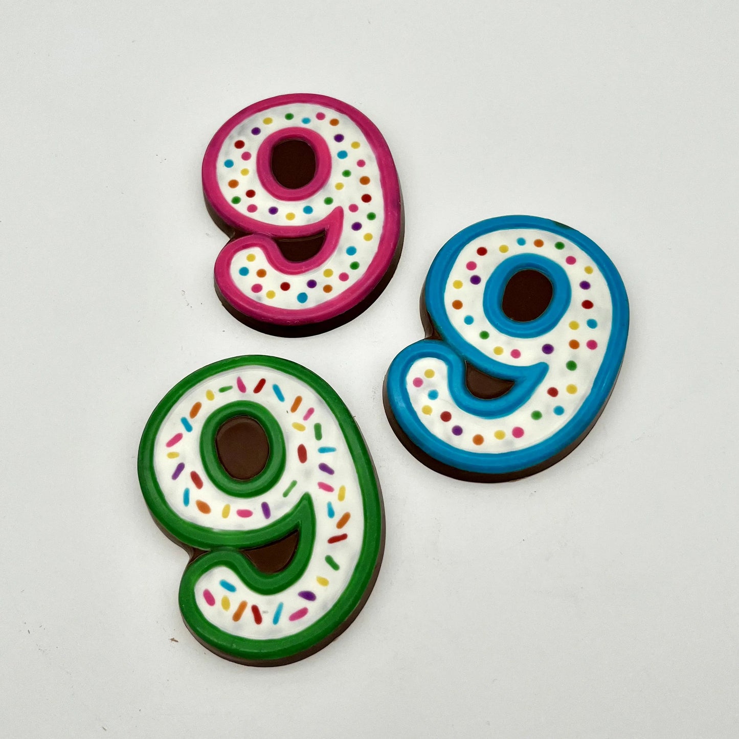 Three colourful number '9' chocolates, hand-painted with multi-colour dots and sprinkles. Displayed on a white background.