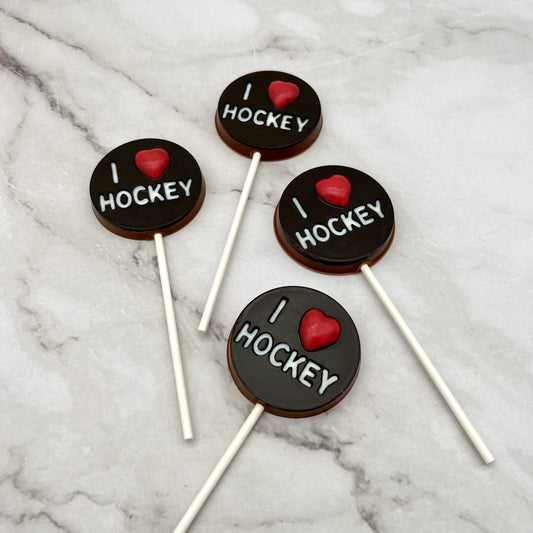 Four 'I ❤️ Hockey' chocolate suckers on a marble surface. White lettering and a red heart on a round black "puck" background.