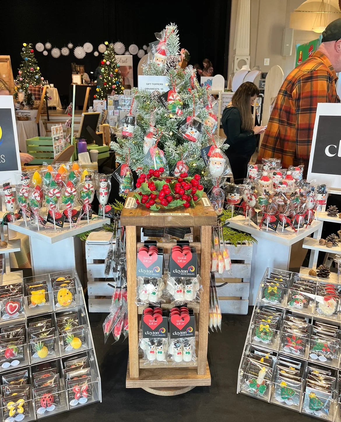 Market display of Christmas-themed chocolates in trays, sucker stands, wooden rotating stand, and a Christmas tree for decoration.